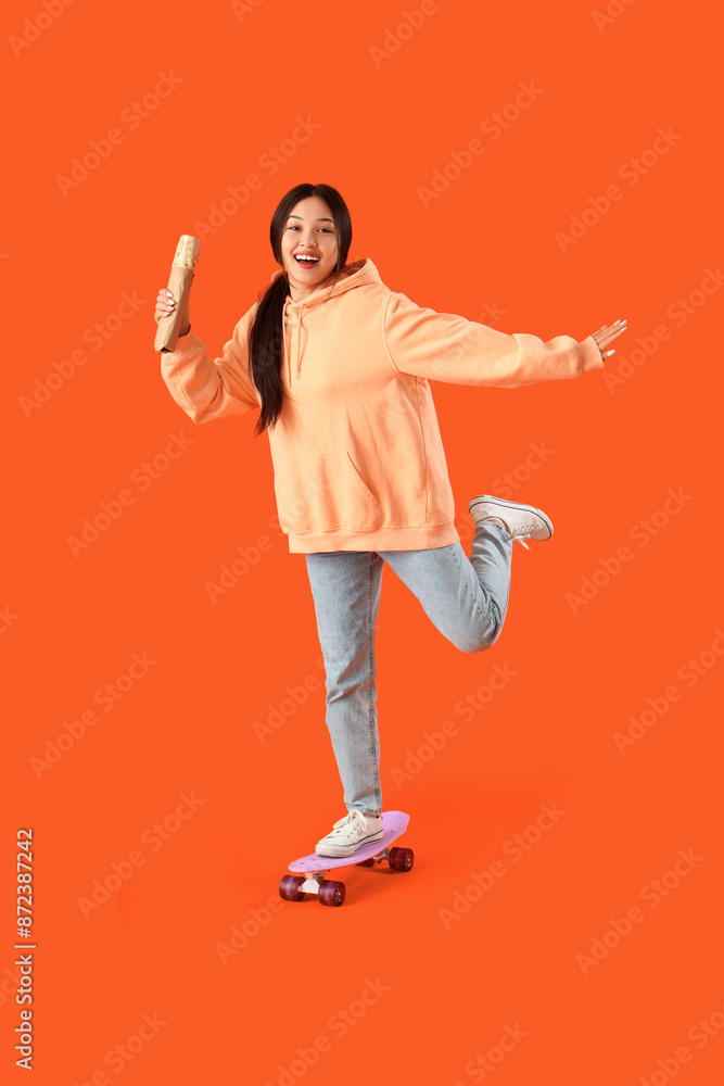 Wall mural Young Asian woman with doner kebab and skateboard on orange background