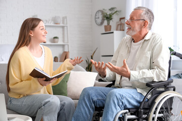Senior man in wheelchair talking to caregiver with book at nursing home