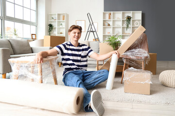 Young man with roll of stretch film and wrapped furniture sitting in room on moving day