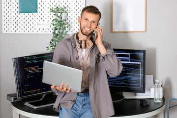 Young bearded programmer with laptop talking by mobile phone in office