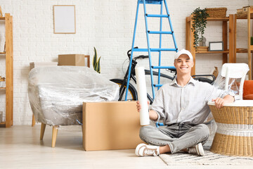 Young man with roll of stretch film sitting on floor at home