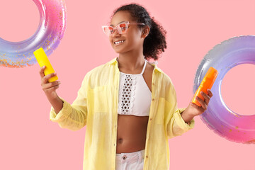 Little African-American girl with bottles of sunscreen cream and swim rings on pink background