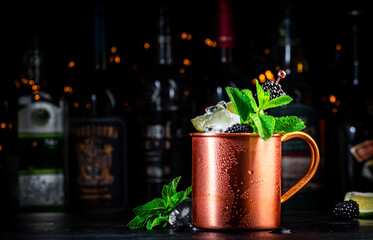 Moscow mule cocktail drink with blackberry in copper mug with lime, ice, ginger beer, vodka and mint. Black bar counter background, bar tools, bottles