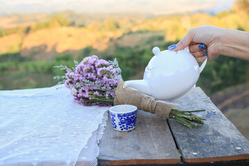 Hand pouring tea from a white teapot with biscuit and violet flower on wooden table landscape...