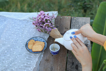 Hand pouring tea from a white teapot with biscuit and violet flower on wooden table landscape...
