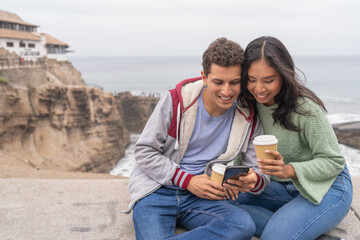Fototapeta premium Couple drinking coffee and using mobile next to the sea