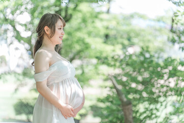 新緑の木漏れ日の中マタニティフォトの撮影をするアジア人妊婦の女性
