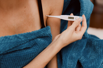 A woman holding an electronic thermometer to her chest wrapped in a blue blanket, wearing a bra and tank top