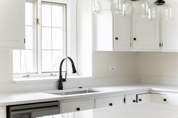 A cozy kitchen faucet detail with white cabinets, black faucet in front of a bright window, and out of focus lights over the marble island.