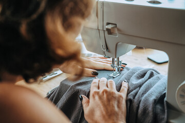 Rear view of a woman seamstress who sits at a sewing machine and sews summer pants from light gray...