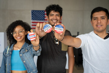 group of Hispanic smiling people holding 2024 Vote election I voted buttons or pins at a US election polling station. In the background, american flag at the background, concept of latin, latino vote