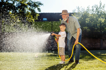 Grandfather is watering lawn with garden hose and his grandson is helping.