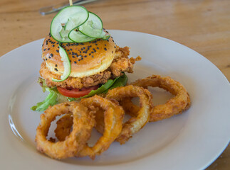chicken burger with cucumber mines on top with breaded onion rings on the side on a white plate, angle view