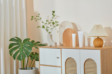 Interior of a bright living room with a wooden cabinet, which containing two blank label cosmetic tubes and many decorative accessories on top, over white wall background. Diagonal angle, blank space