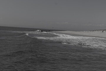 Beautiful image of the Jersey shore. The white froth of the waves crashing in to the beautiful brown sand of the beach. A small jetty with black rocks can be seen. The blue sky has some white clouds.