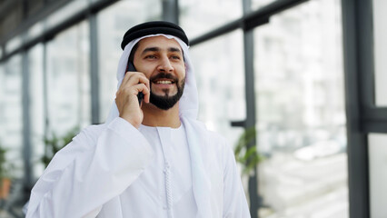 Professional Arab UAE Emirati man using smartphone on call. Happy Middle Eastern national wearing kandura at a business corporate location