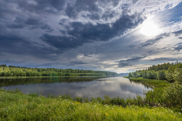 A serene lake reflecting the dramatic hues of a stormy sky, surrounded by vibrant green trees and wildflowers.