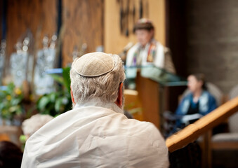 An elderly man with white hair wearing a jewish prayer shawl and a yamaka with a bar mitzvah boy at a podium in the distance at a synagouge