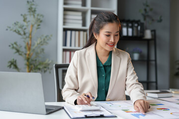 Asian business woman happily recording data on laptop Details from the graph Business income charts in real estate, taxes, management, technology marketing online in the office.