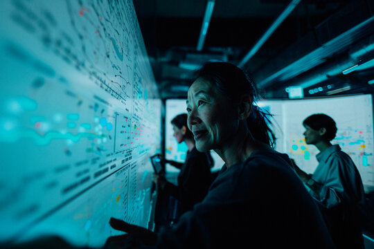 A team of individuals working together in a dark room, analyzing complex data on screens and large boards, showcasing collaborative effort and focus in a tech-driven environment.