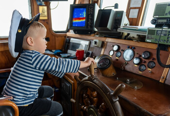 Young boy playing captain in wheelhouse, exploring navigation and adventure. Baby is dressed in a sailor hat and striped shirt, adding to the nautical theme. High quality photo . High quality photo © Maryia