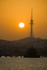 Sunset over Qingdao with the TV tower and traditional pavilion silhouetted against an orange sky