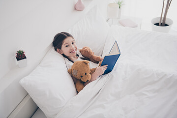 Portrait of adorable small girl read book teddy bear toy wear pajama comfy bed bedroom apartment indoors