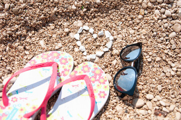 Beach season. Sunglasses and flip-flops lying on a rocky beach with a heart made of stones