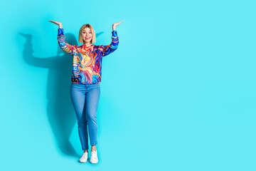Full size photo of positive girl dressed print shirt raising hands up presenting object empty space isolated on blue color background