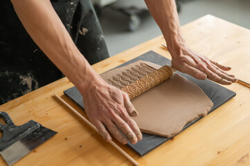 Close-up of a potter's hands rolling out clay using a rolling pin with patterns. 