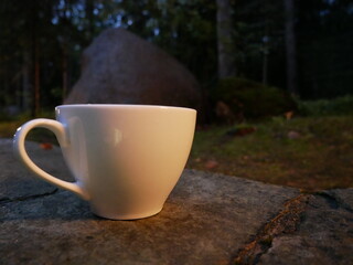 A white porcelain cup on a background of trees on a summer night. Tea party in the evening outside on the street. Night tea drinking in the dark outdoors.