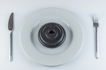Round white pottery dish, with a black heavy weight in the middle. Fork on the right and knife on the left, with white background. Useful for concepts like diet, heavy food, indigest.