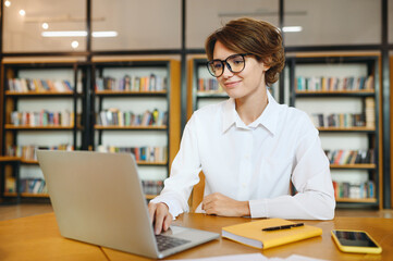 Young smiling happy successful employee IT business woman wear white shirt casual clothes glasses hold use work on laptop pc computer surfing internet sit at office desk. Achievement career concept.