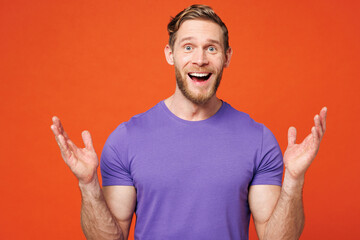 Young surprised excited jubilant shocked happy cool man he wear purple t-shirt casual clothes looking camera spreading hands isolated on red orange wall background studio portrait. Lifestyle concept.