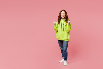 Full body young ginger woman she wear green hoody casual clothes doing winner gesture celebrate clench fists say yes isolated on plain pastel light pink background studio portrait. Lifestyle concept.