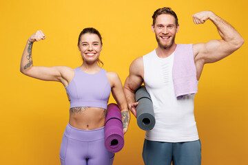 Young strong fitness trainer instructor sporty two man woman wear blue clothes spend time in home gym hold yoga mat caremat show muscles isolated on plain yellow background. Workout sport fit concept.