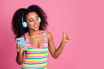 Portrait of attractive girl in headphones hold smartphone look indicating at discount empty space isolated on pink color background