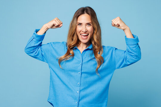 Young strong happy woman wear shirt casual clothes showing biceps muscles on hand demonstrating strength power isolated on plain pastel light blue cyan background studio portrait. Lifestyle concept.