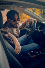 A stylish man in glasses sits behind the wheel, holding the steering wheel with the sun shining, emphasizing the use of sunglasses for driving safety against glare.