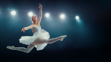 Graceful Ballet Dancing Ballerina in White Tutu Performs Stunning Grand Jeté Under Stage Lights, Her Elegance and Athleticism in Captivating Ballet Performance. © Gorodenkoff