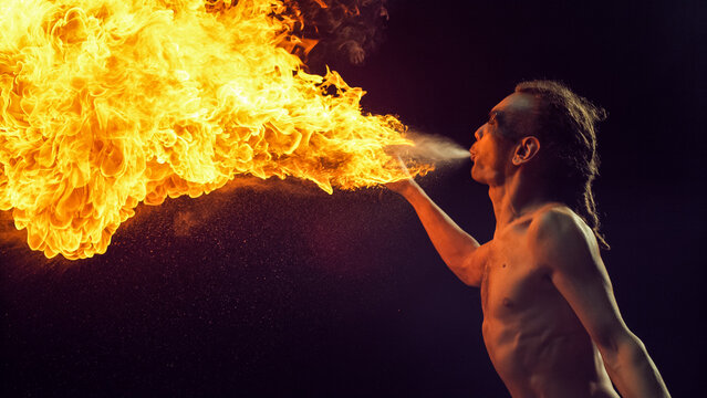 Male Fire Performer With Dreadlocks Skillfully Exhales Massive Fireball, Creating Spectacular Display of Flames and Sparks Against Dark Background, Cinematic Intensity and Artistry of Fire Breathing. - Powered by Adobe