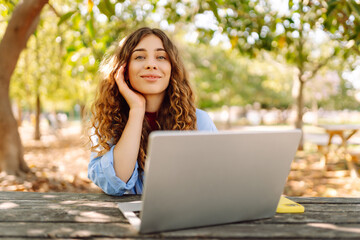 Young woman sits in park with laptop. Remote work or education.  Freelance, online course.