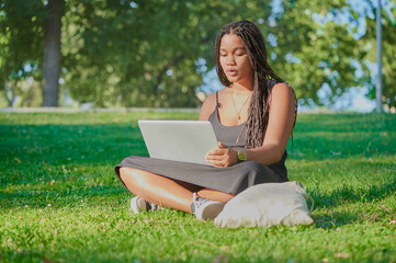 Surprised biracial teenage girl with braided hair checks information on the internet on her laptop which she uses remotely from a park on a sunny day.