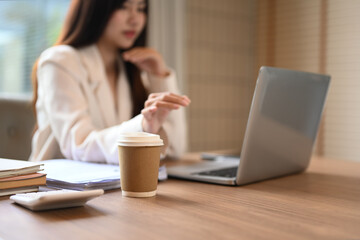 A paper cup of coffee on wooden table with calculator and paperwork, businesswoman sitting in background