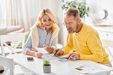 A mature loving couple in cozy homewear sit together at a table, enjoying a quiet moment together in their kitchen at home.