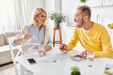 A mature man and woman in cozy homewear sitting at a table, focused on using a calculator for financial calculations.