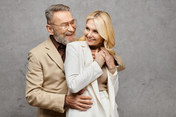 A mature man and woman, elegantly dressed, standing close together in a loving embrace on a gray backdrop.