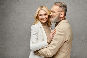 A mature man and woman in elegant attire embrace each other tenderly against a gray backdrop.