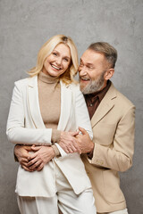 An elegant mature couple in debonair attires sharing a warm and tender embrace on a gray backdrop.