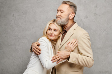 A mature loving couple in debonair attire warmly embrace each other in a graceful pose against a gray backdrop.